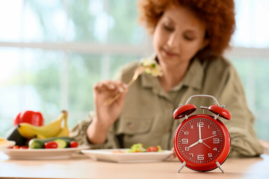 Red alarm clock on a table with a woman eating a healthy meal in the background, timed eating and diet