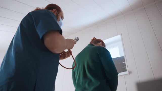 Paramedic home visit checking back with stethoscope, male clinician in blue scrubs and mask examines standing patient near window inside bright house, attentive posture and gentle reassurance