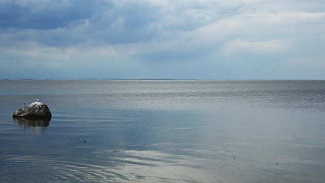 View across a large calm lake with terns on a rock and a sky full of blueish clouds