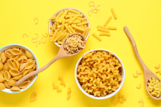 Bowls and wooden spoons with different types of tasty raw pasta on yellow background