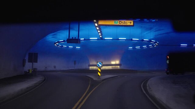 Driving through a Norwegian road tunnel featuring a distinctive underground roundabout and atmospheric blue lighting. The illuminated tunnel interior, road signs and passing traffic showcase Norway's 