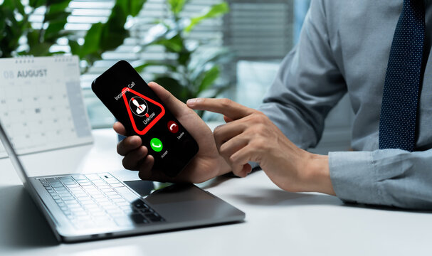 Businessman receives a suspicious unknown caller alert on smartphone beside a laptop, highlighting phone scam, caller fraud, cybersecurity risk, phishing, and mobile communication security.