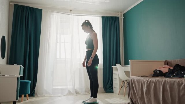 Home bedroom woman steps onto scale for check. Sunlit bedroom with teal wall and scattered clothes on bed, young woman in workout top and leggings approaches teal digital scale on rug with focused