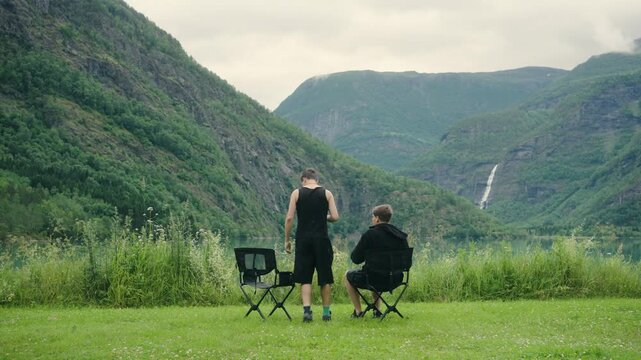 Cinematic footage of two male travelers relaxing in folding chairs with hot drinks beside Eidsvatnet Lake near Skjolden in western Norway. They sit peacefully overlooking the emerald lake, surrounded 