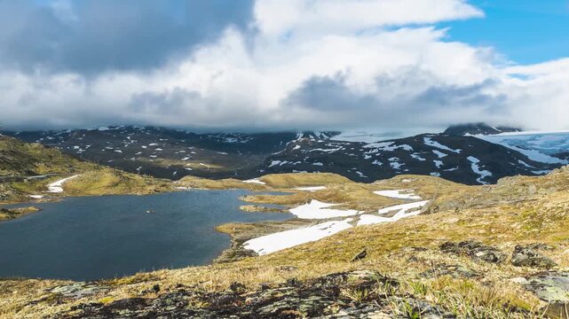 Cinematic timelapse footage captured in Breheimen National Park, Norway, showcasing a rugged alpine landscape beneath fast-moving clouds. Rocky mountain terrain is interspersed with patches of snow