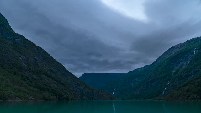 Cinematic timelapse footage capturing dark storm clouds racing across the sky above Eidsvatnet Lake in western Norway, near the inner reaches of Sognefjord. The composition features calm lake water