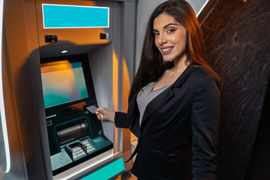 A smiling young woman in a black blazer looks toward the camera while inserting a card into an ATM at night