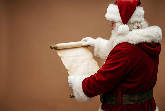 Santa claus in a classic red suit reading a long paper scroll against a solid brown studio wall