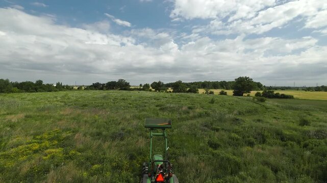 Wide aerial tracking shot flying forward over a compact utility tractor pulling a rotary brush hog mower to cut down tall weeds and clear a path across a large field.