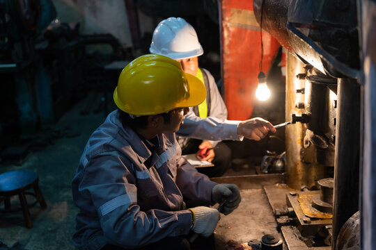 Senior engineer pointing and instructing technician wearing yellow hardhat during machinery maintenance in factory.