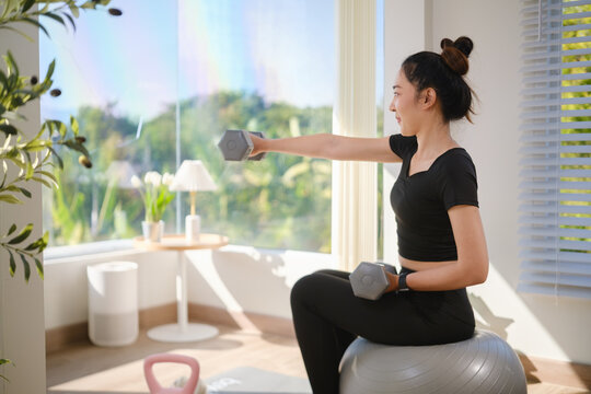 Young Asian woman performing a seated dumbbell exercise on a stability ball at home.