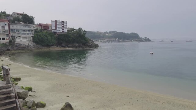 Wide angle view of a deserted sandy beach, calm water, and residential seaside buildings under soft, overcast daylight for serene, moody footage.