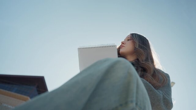 Pretty asian poet reading notebook on concrete ledge, low angle composition with knees foreground, soft sun halo behind hair, quiet reflective mood, urban backdrop and casual coat