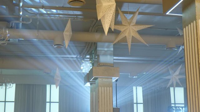 Low-angle view of industrial ceiling with exposed pipes, hanging white paper stars, structural columns, and dramatic light rays streaming through windows creating festive banquet atmosphere 4k video