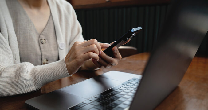 Woman using a smartphone while working on a laptop at a wooden desk, representing digital lifestyle, mobile communication, technology and modern productivity.