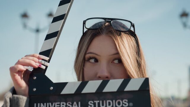 Stylish woman with filming equipment. Glamorous woman wearing sunglasses while holding film slate outside. Beautiful actress standing outdoors with script and sunglasses in urban environment