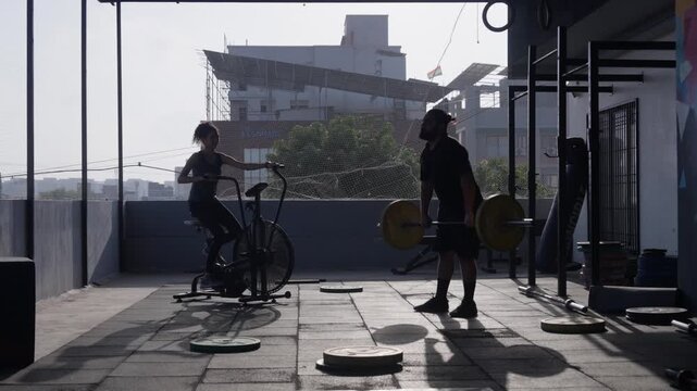 South Asian female riding an air bike while male athlete performs barbell cleans during a workout at an open-air rooftop gym. Wide shot with lateral camera movement. Collection: WORKOUTSERIES2026.