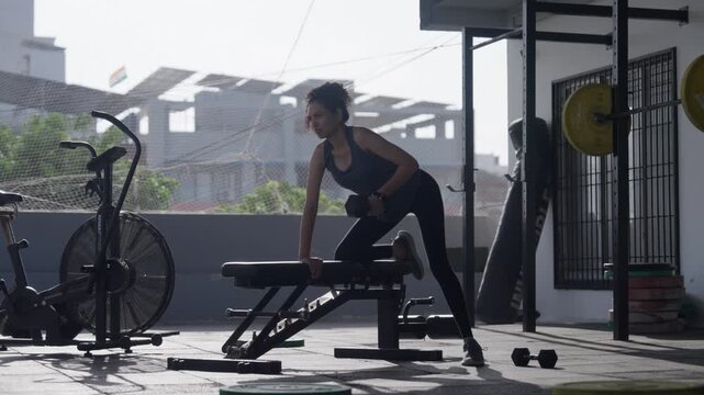 South Asian female athlete performing single-arm dumbbell rows supported on a bench at an open-air rooftop gym. Full shot with static camera angle. Collection: WORKOUTSERIES2026.