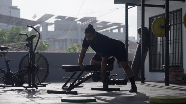 Bearded South Asian male athlete performing single-arm dumbbell rows supported on a bench at an open-air rooftop gym. Full shot with static camera angle. Collection: WORKOUTSERIES2026.