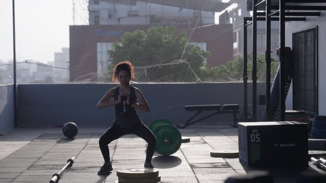 South Asian female athlete doing dumbbell goblet squats at an open-air rooftop gym. Wide with lateral camera movement showcasing controlled squat technique. Collection: WORKOUTSERIES2026.