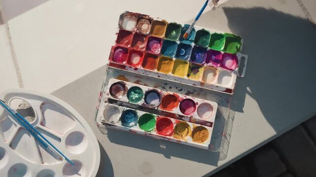 Pretty watercolor palette on sunlit tabletop, colorful pans arranged, round mixing tray with clean wells, slender brushes beside palette, warm natural light casting soft shadows, minimal serene art