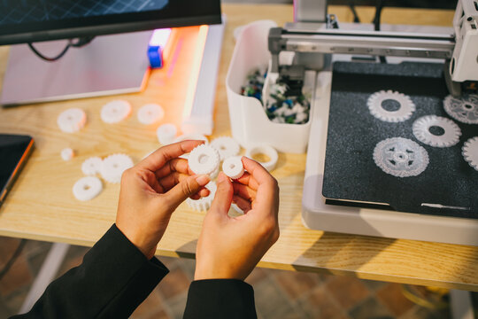 Hands hold white 3d printed gears beside a desktop printer in a workshop, prototyping, design and manufacturing.