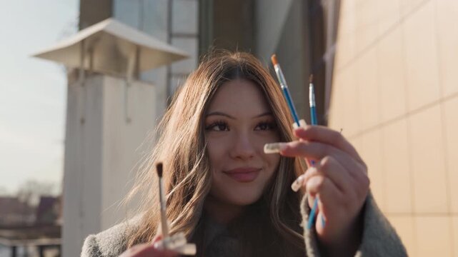 Woman with brushes glows warmly. Charming rooftop artist with loose hair under warm glow. Portrait of woman in coat smiling with sunlight catching her flowing hair outside urban background