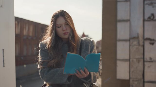 Photo of woman reading on rooftop during sunset. Closeup of person journal reading against backlit urban background. Portrait of woman engrossed in journal while shaded by warm sunset light