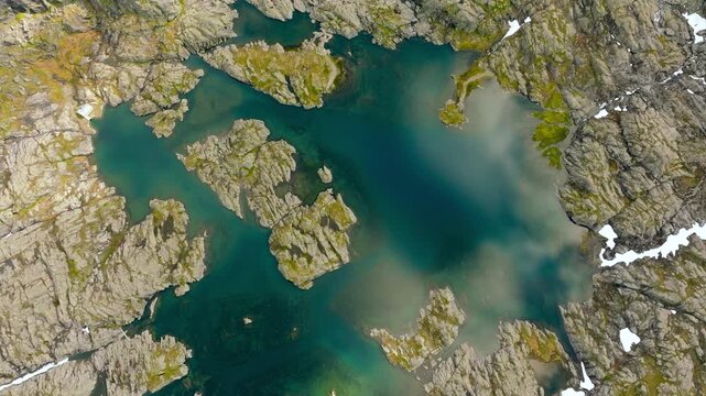 Aerial drone footage looking straight down over a small high-altitude mountain lake surrounded by rugged rocky terrain and patches of snow along the Trolltunga hiking route in Norway. The drone moves 