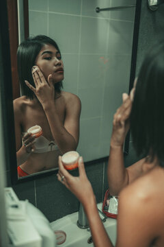Asian woman looks into a bathroom mirror while applying facial skincare cream with a cosmetic pad, showing everyday beauty, grooming, skin treatment, and self-care.