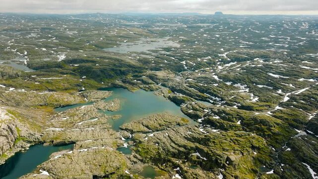 Aerial drone footage of the rugged high mountain plateau surrounding Trolltunga in Norway. The expansive landscape features bare rocky terrain, numerous mountain lakes, patches of lingering snow 