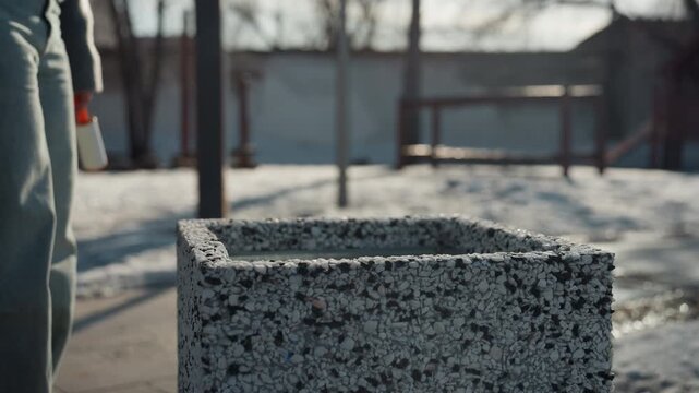 Pretty granite planter beside snowy sidewalk pedestrian drops red lighter into planter closeup of hand and textured stone muted winter light shallow depth of field cinematic quiet mood urban bench