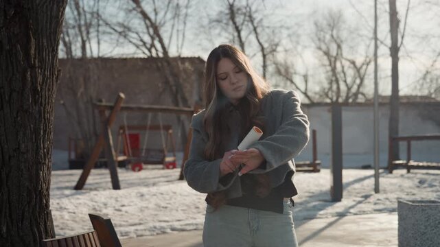 Pretty woman rolling lint roller on sleeve, sunlit playground and snowy ground backdrop, focused routine and steady hand motion, commuter or barista prepping uniform before shift, candid streetstyle