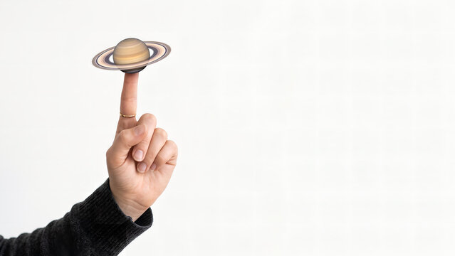 Surreal conceptual photo of a human hand balancing planet Saturn on a fingertip over bright white minimalist background.