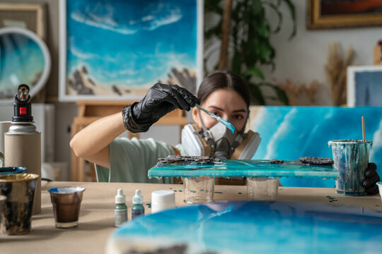 Woman crafter in respirator works with epoxy resin using canvas and spatula at table in studio, professional female artist adding liquid resin on painting, creating fluid art masterpiece in workshop.