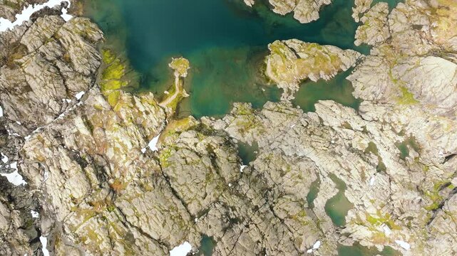 Aerial drone footage looking straight down over a small high-altitude mountain lake surrounded by rugged rocky terrain and patches of snow along the Trolltunga hiking route in Norway. The drone moves 