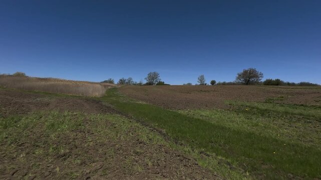 Aerial view starting close to the ground before ascending smoothly upwards to reveal rolling agricultural fields with patchy green growth and raw tilled soil under a clear sky.