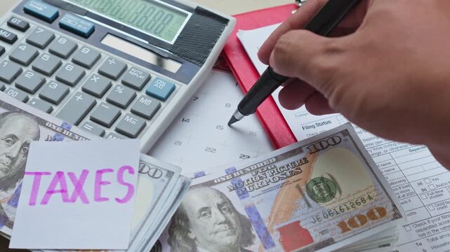 Hand marks date on calendar next to tax form, calculator, cash, and taxes note
