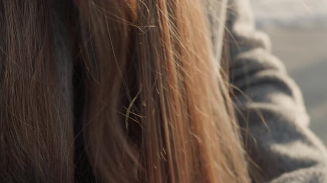 Pretty asian woman brushing long hair on bench, park visitor detangling strands in gray coat, soft sunlight, winter scene, closeup fingers smoothing locks, quiet reflective mood.