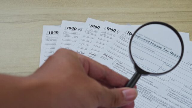 Hand uses magnifying glass to inspect U.S. individual income tax return 1040 forms on desk