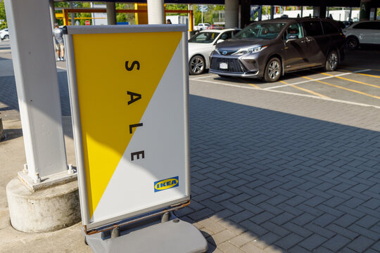 A yellow and white IKEA SALE sandwich board stands in the covered parking area, its small blue logo below the diagonal lettering, with vehicles parked behind.