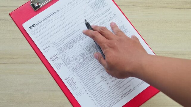 Hand fills out U.S. individual income tax return 1040 form on red clipboard