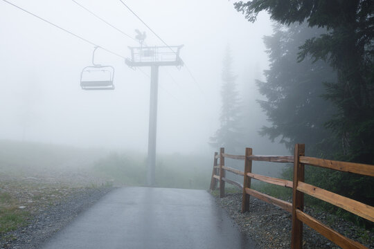 A chairlift tower and empty chair rise into dense fog above a wet paved path on Grouse Mountain, with a rustic split-rail fence and evergreens along the misty slope.