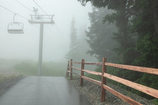 An empty chairlift chair hangs from a tower shrouded in fog on Grouse Mountain, where a wooden split-rail fence leads along a wet path into misty coastal forest.
