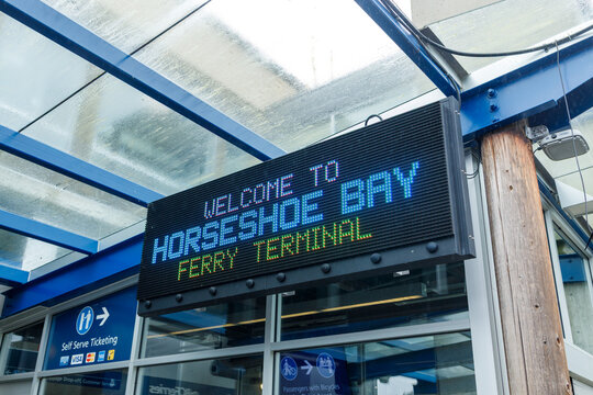 A digital LED sign reads Welcome to Horseshoe Bay Ferry Terminal beneath the blue steel and glass canopy of the BC Ferries terminal entrance.
