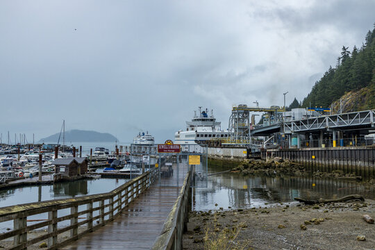 A wet wooden boardwalk runs past a Sewell's Marina East Moorage sign toward the BC Ferries terminal at Horseshoe Bay, ferries berthed under a grey sky.