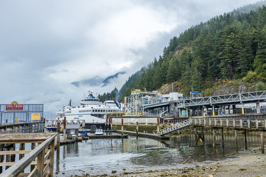 A BC Ferries vessel rests at the terminal as cloud lifts off the green forested mountain, gangways reaching to floats past the gated Sewell's Marina boardwalk.