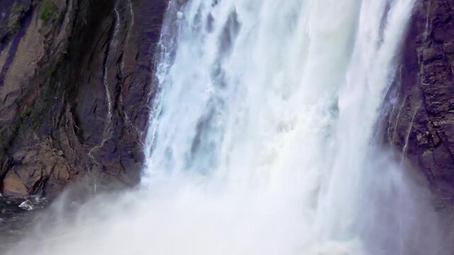 Panning low angle view of powerful Montmorency Falls cascading down cliffs under a bridge in Quebec City, Quebec, Canada.