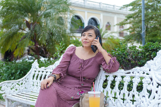 Woman in Purple Dress Talking on Phone on White Bench in Tropical Plaza