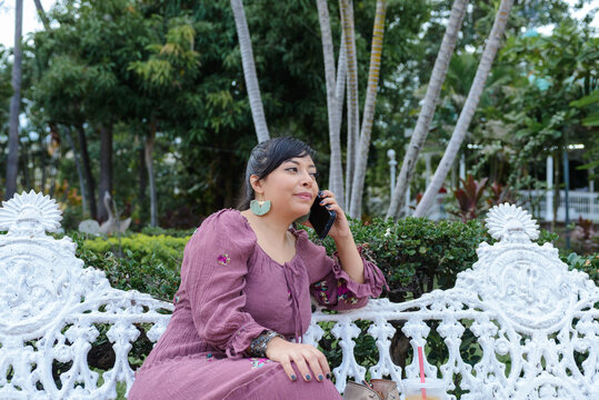 Woman in Purple Dress Talking on Phone on White Bench in Tropical Plaza